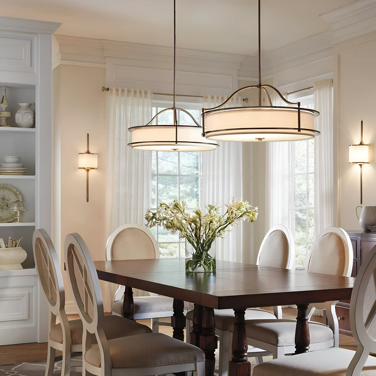 A dining room showcasing a square wooden table is surrounded by six white cushioned chairs. Elegance and warmth are added by two Emory 4 Light Duo-Mount Chandeliers in Olde Bronze hanging above. A vase with flowers adorns the table, while large windows with sheer curtains and subtle wall lighting complete the ambiance of the room.