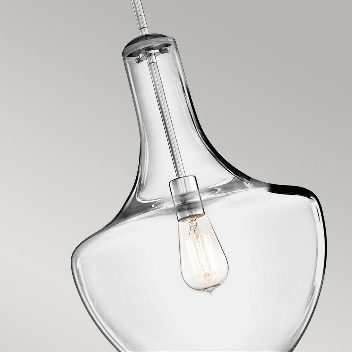 A detailed view of the Everly 1 Light Medium Pendant in Chrome showcases a vintage-style LED filament bulb inside its clear glass shade, set against a simple gray backdrop. The illuminated bulb emits a warm glow through the transparent glass.