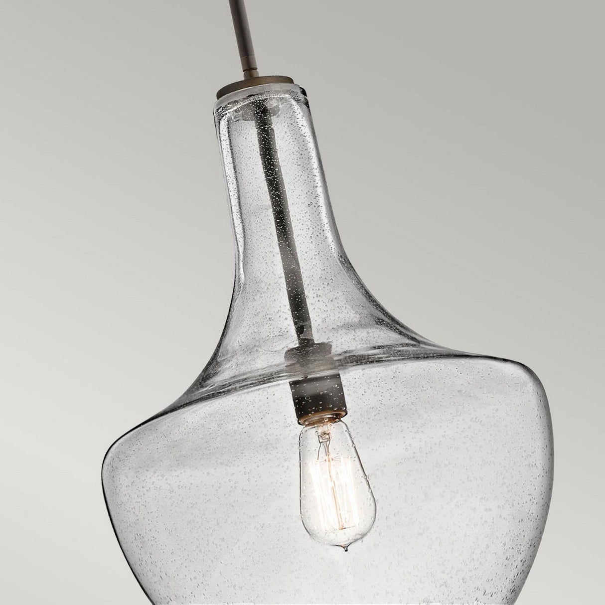 A close-up of the Everly 1 Light Medium Pendant - Olde Bronze showcases a modern design, featuring a large clear seeded glass shade that beautifully displays the visible light bulb. Suspended from a cord against a plain gray background, its textured glass delivers an elegant bubble-like appearance.