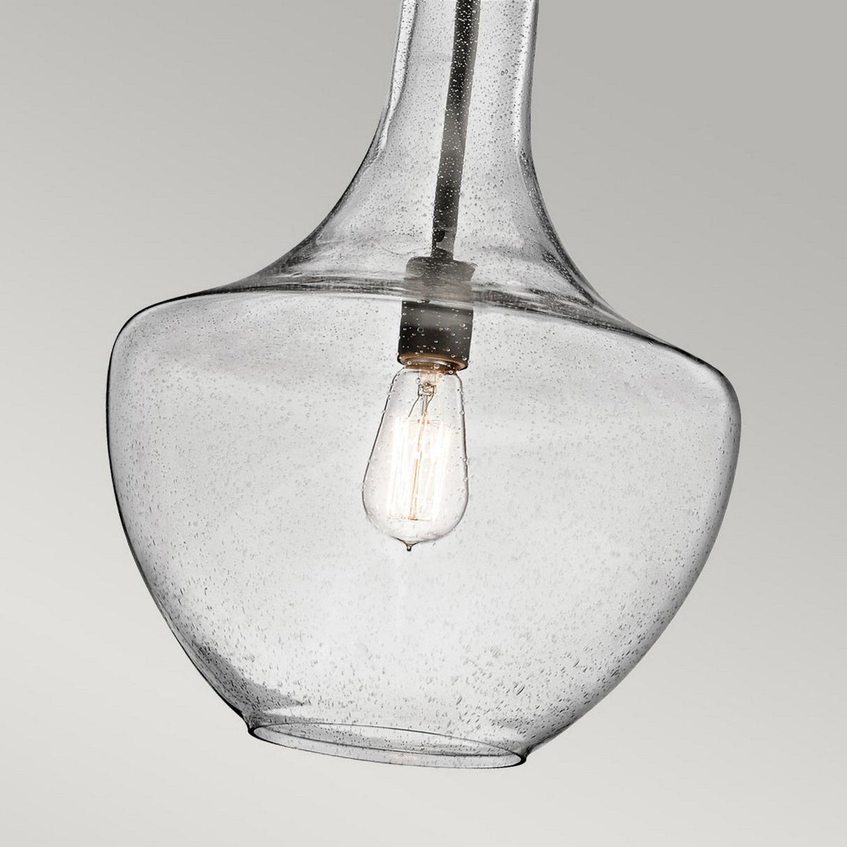 The Everly 1 Light Medium Pendant - Olde Bronze showcases its clear seeded glass housing a visible filament bulb, set against a plain gray backdrop. The bulb's understated vintage-style design harmonizes beautifully with the contemporary elegance of the glass and its refined Olde Bronze finish.