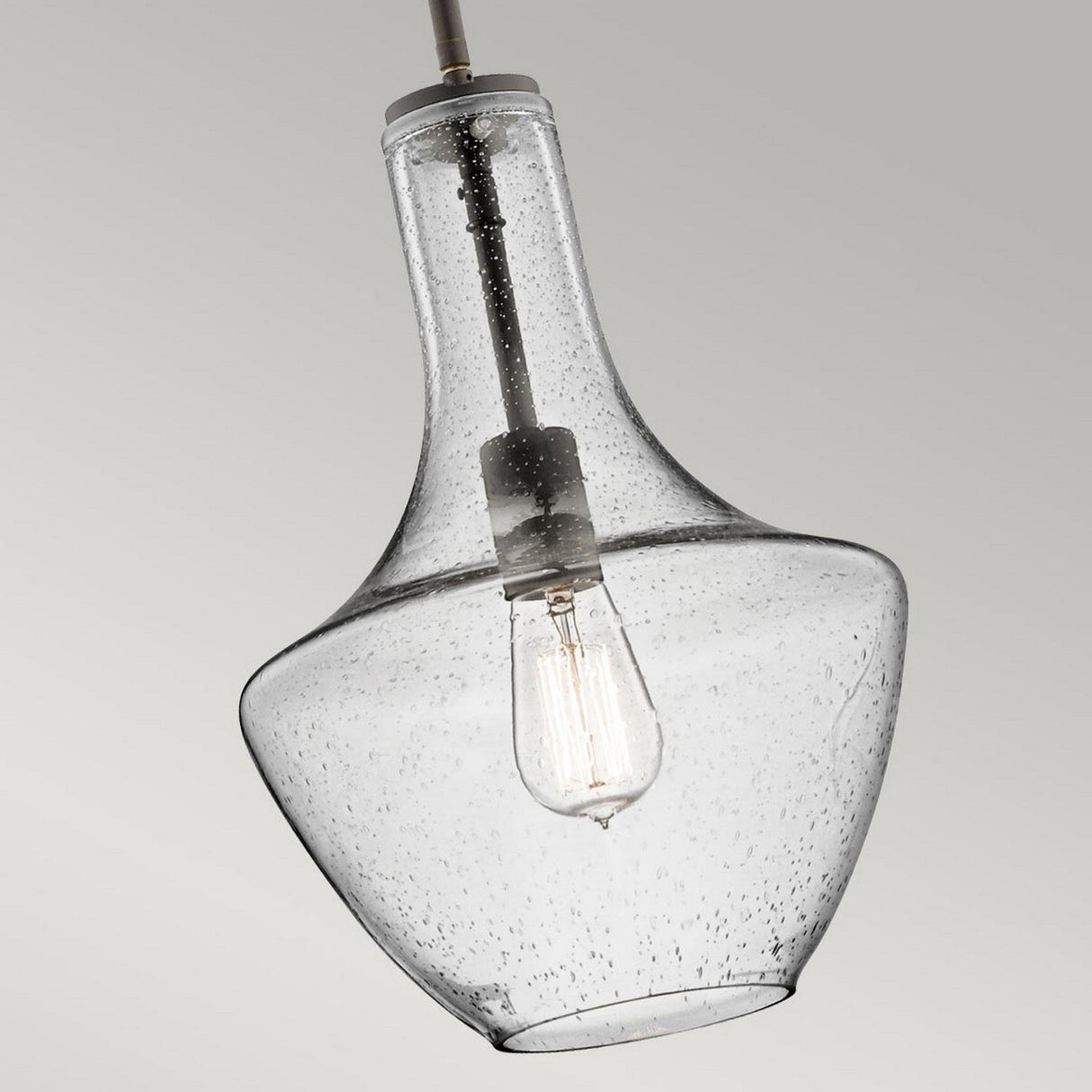 An Everly 1 Light Small Pendant in Olde Bronze hangs from the ceiling, featuring a teardrop-shaped clear seeded glass that showcases its charming vintage-style LED filament bulb against a light gray background. The glass has a subtle texture with tiny bubbles scattered throughout, enhancing the elegance of its Olde Bronze finish.