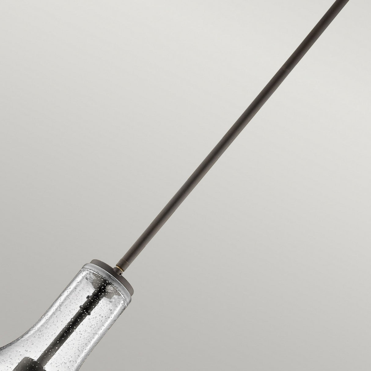 A close-up of the Everly 1 Light Small Pendant in Olde Bronze showcases its clear, seeded glass shade. The long, slender rod stands out gracefully against a neutral gray background, accentuating the pendant's sleek and minimalist design.