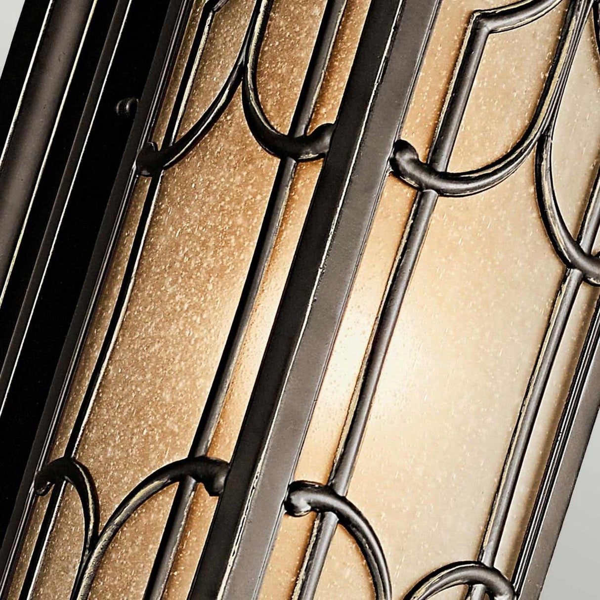 A close-up of the elegant Franceasi Outdoor 1 Light Medium Wall Lantern in Olde Bronze, showcasing its frosted glass with a textured finish evocative of European castle windows. It's encased in a decorative black metal frame featuring intricate geometric patterns that softly illuminate the glass, highlighting the design details.
