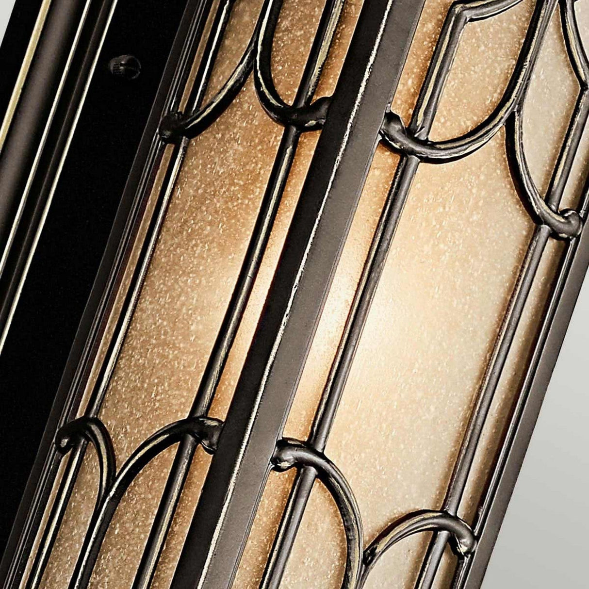 Close-up of the Franceasi Outdoor 1 Light Small Wall Lantern in Olde Bronze, showcasing intricate metalwork and frosted, amber-tinted glass panels that emit a warm, ambient glow. The design includes geometric patterns with metallic frames.