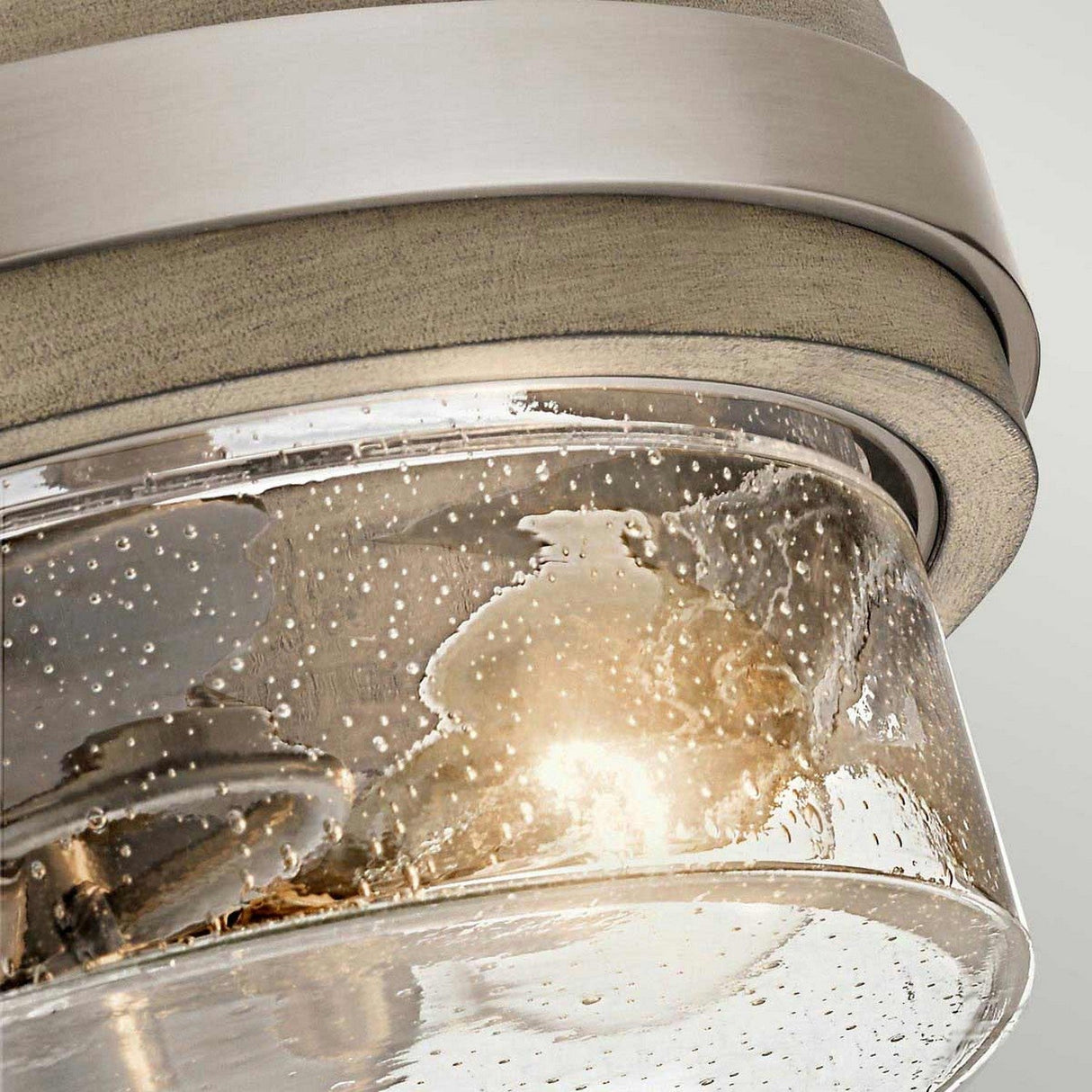 A detailed view of the Grand Bank 2 Light Semi Flush in Antique Grey showcases its clear glass cover adorned with bubble patterns and complemented by Antique Grey metal accents. The warm glow from inside enhances its semi-flush design, blending modern style with vintage industrial appeal.