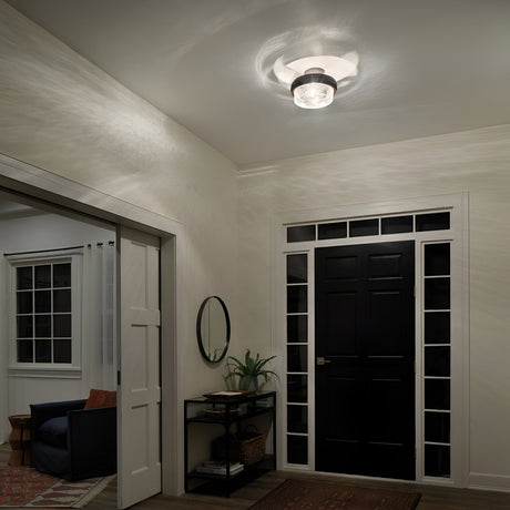 A modern foyer with a black front door framed by glass panels exudes vintage industrial charm. The Grand Bank 2 Light Semi Flush in antique grey casts soft shadows. A round mirror and plant rest on a small table beside the door, leading to an adjacent room featuring a chair with pillows in antique grey.