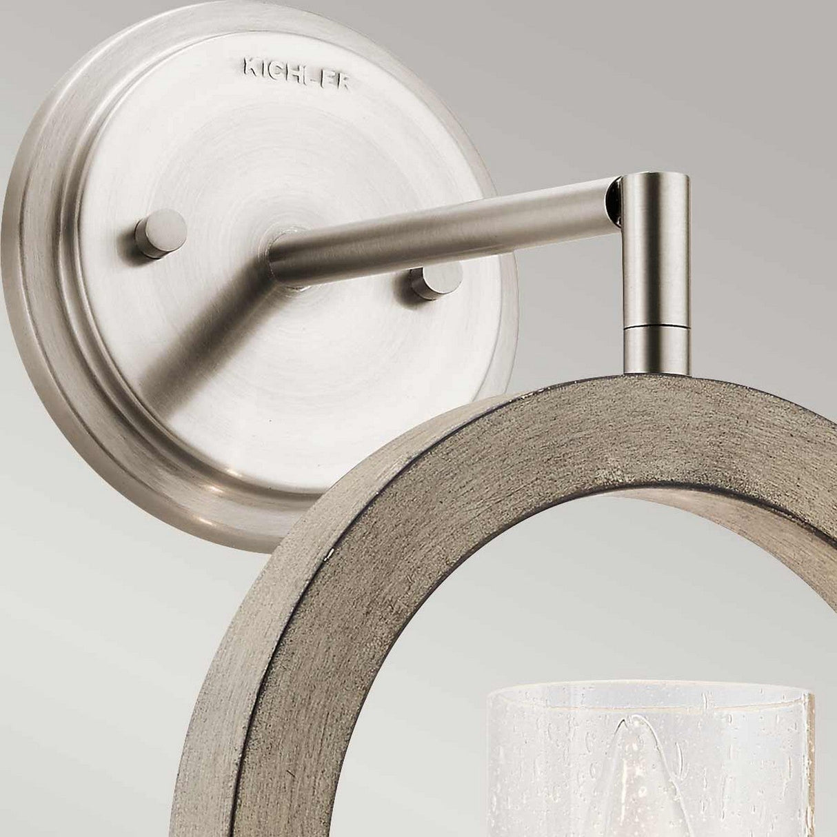 Close-up of the Grand Bank 1 Light Wall Light in Antique Grey, showcasing its modern design. The fixture includes a brushed metal finish with a round backplate and an angular arm supporting a circular, textured frame that holds a cylindrical, frosted glass light cover.