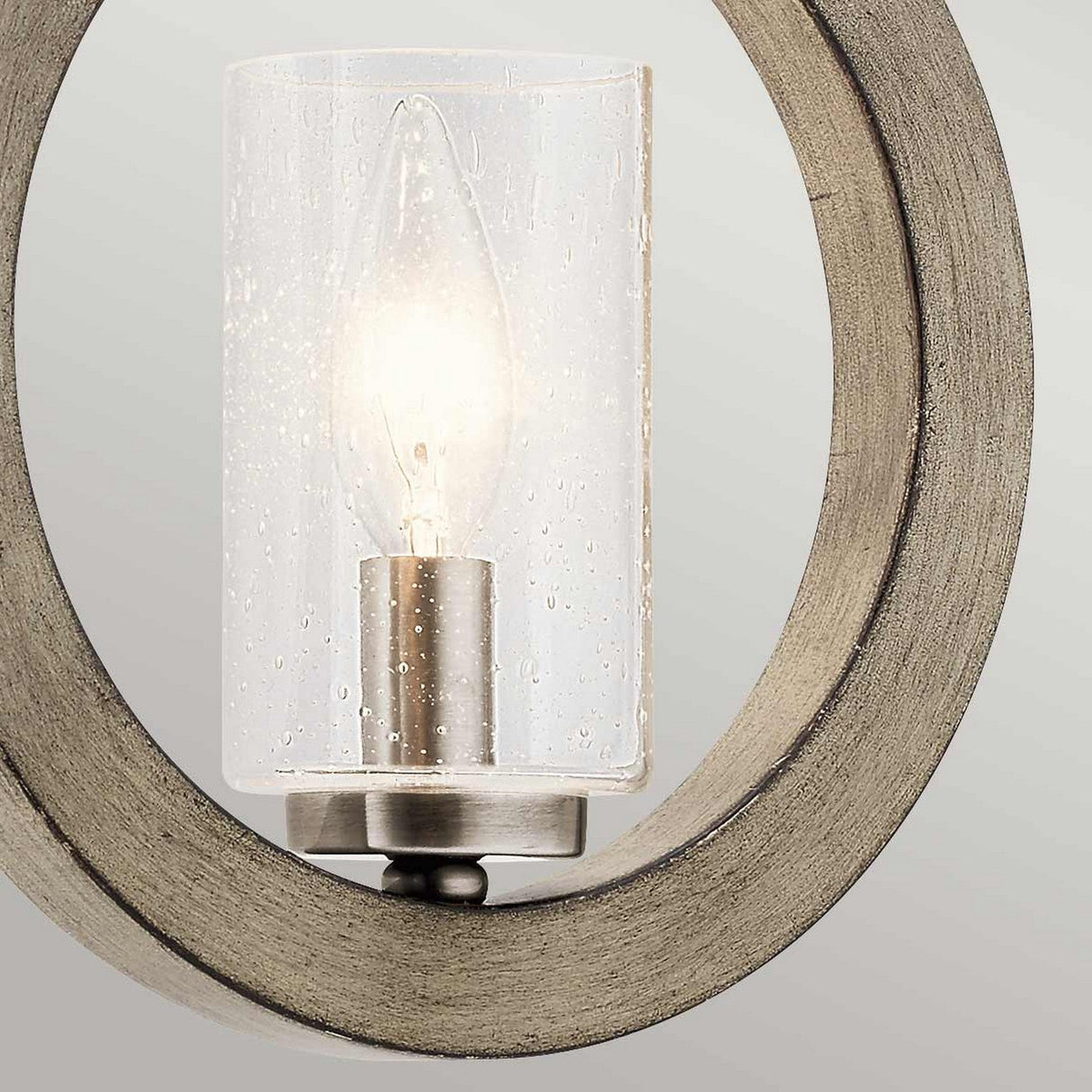 A close-up of the Grand Bank 1 Light Wall Light in Antique Grey showcases its rustic charm. The view highlights a cylindrical, clear glass cover with a textured surface and reveals a lit candle-style bulb housed within a circular, metallic beige frame. This elegant light is set against a light gray background, perfectly blending modern style with antique nuances.