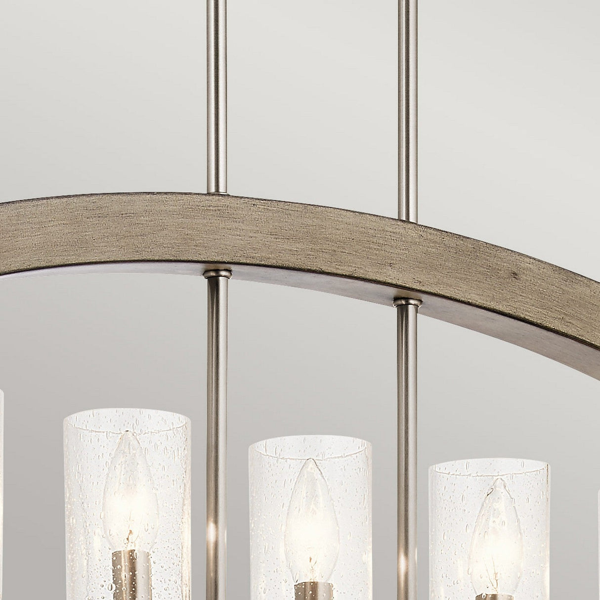 A close-up of the Grand Bank 5 Light Chandelier in antique grey showcases its gracefully curved metal frame and multiple cylindrical glass shades. The shades encase glowing light bulbs, with visible wires extending upward. The neutral light gray background perfectly highlights its timeless elegance.