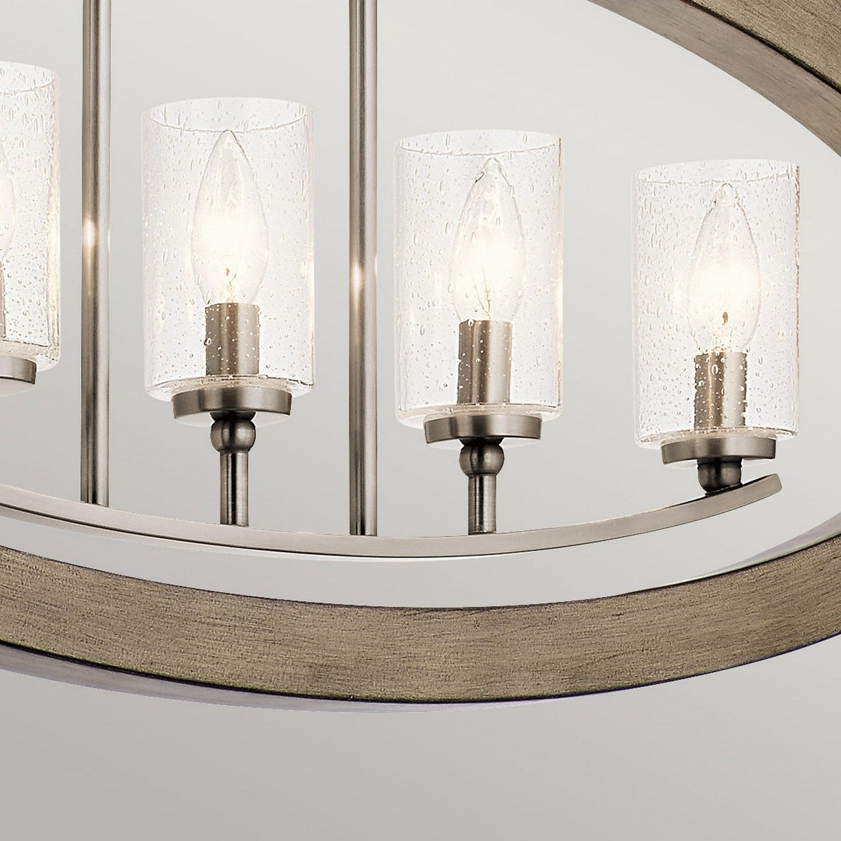 The Grand Bank 5 Light Chandelier in Antique Grey showcases three modern, cylindrical clear glass shades with visible candle-style bulbs. It is elegantly mounted on an antique grey frame accented with wood, featuring soft curves for a contemporary design.