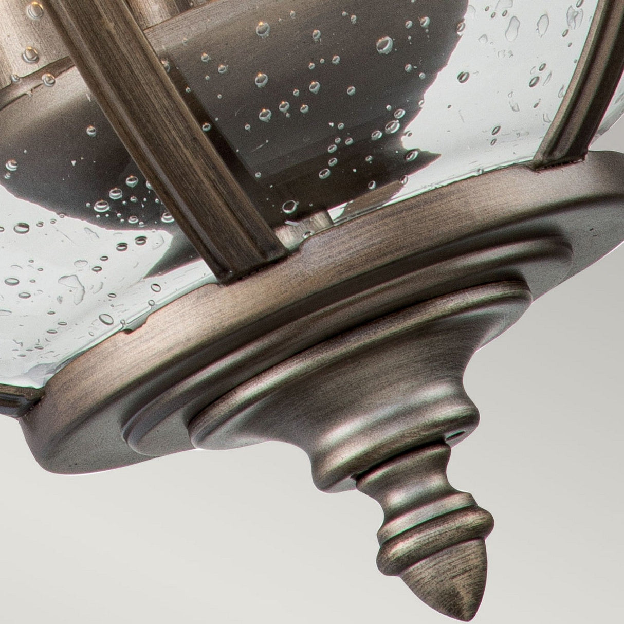 Close-up of the Halleron Outdoor 3 Light Wall Lantern in burnished bronze, featuring a clear glass cover adorned with small water droplet patterns. The lantern radiates Victorian elegance with its decorative finial and metal bars encasing the glass.