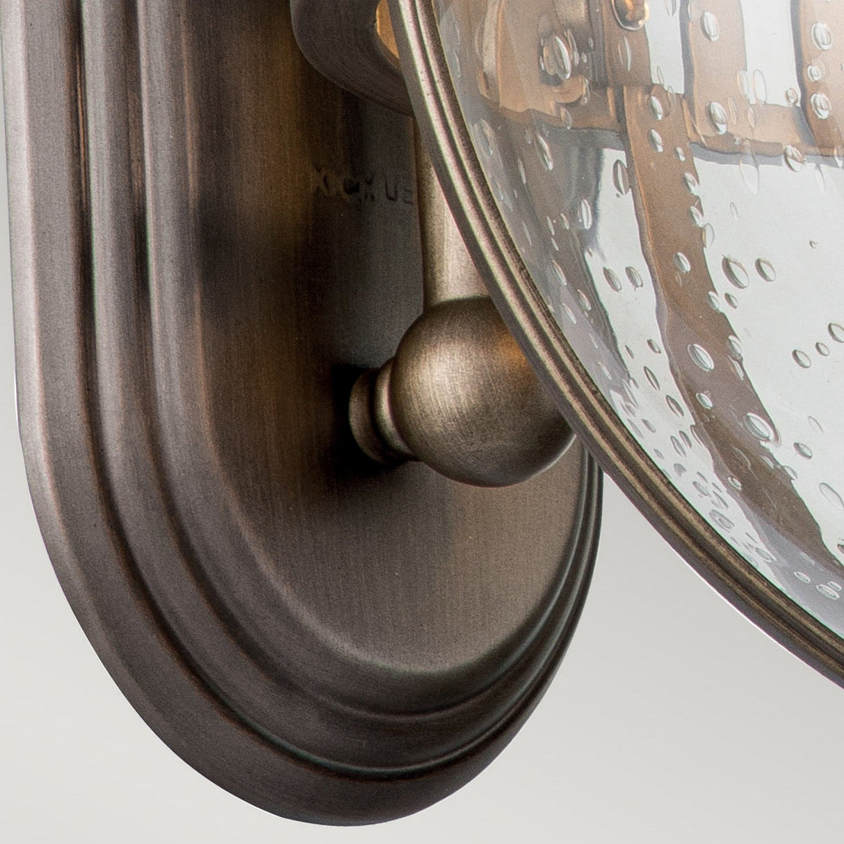 A detailed view of the Halleron Outdoor 3 Light Wall Lantern reveals its oval, burnished bronze backplate and clear, speckled glass shade. This fixture embodies Victorian elegance, featuring a design that encloses bulbs within the glass—a perfect choice for outdoor lighting.