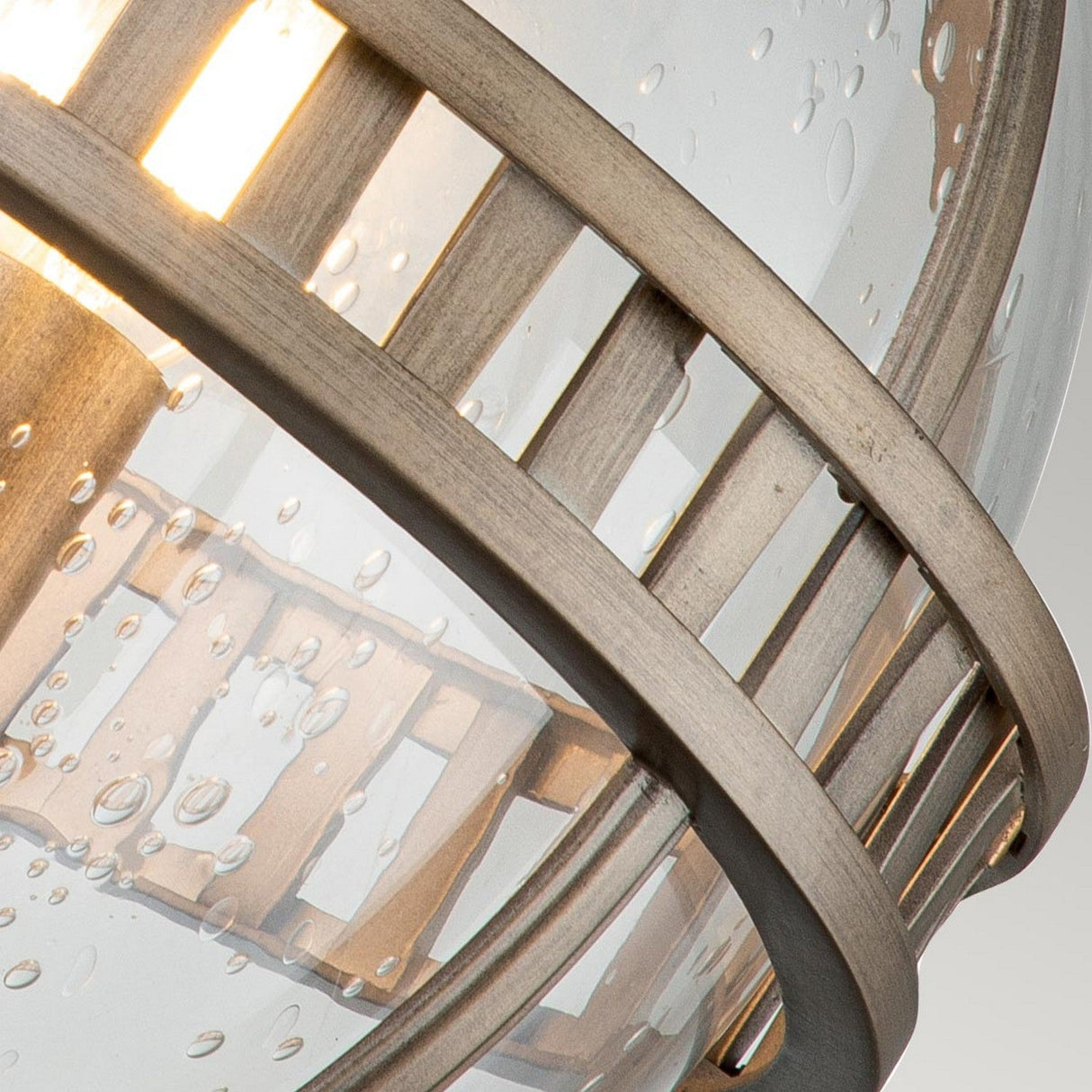 Close-up of the Halleron Outdoor 3 Light Wall Lantern, which showcases a clear glass globe with small bubbles inside. Encased in elegant burnished bronze bands reminiscent of Victorian style, it provides warm outdoor lighting from within, achieving a harmonious blend of classic charm and modern design.