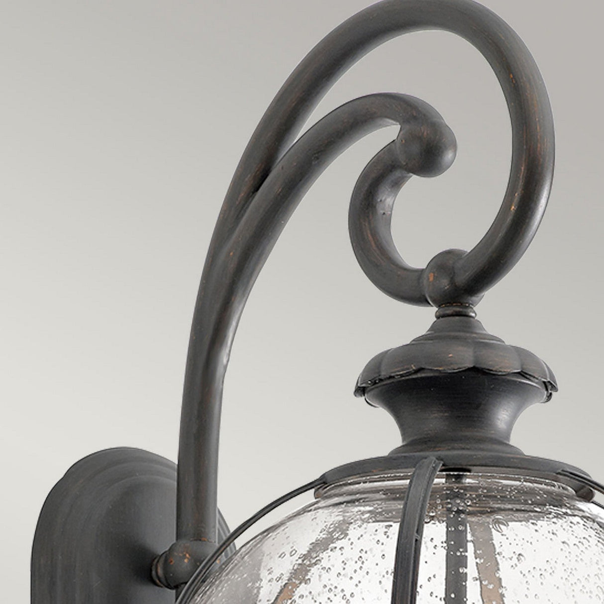 Close-up of the Halleron Outdoor 3 Light Wall Lantern, showcasing its vintage-style charm with a dark metal finish. This lantern features a decorative scroll arm and a clear glass shade with a bubbled texture, set against a simple light grey background.