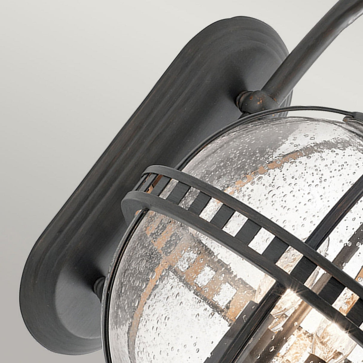 Close-up of the Halleron Outdoor 3 Light Wall Lantern in grey, showcasing a vintage-inspired design with a black metal frame that embodies Victorian sophistication. It exudes classic charm through its elegantly curved arm and clear, textured glass shade. The illumination inside casts a warm glow, and the fixture is mounted on a straightforward rectangular base.