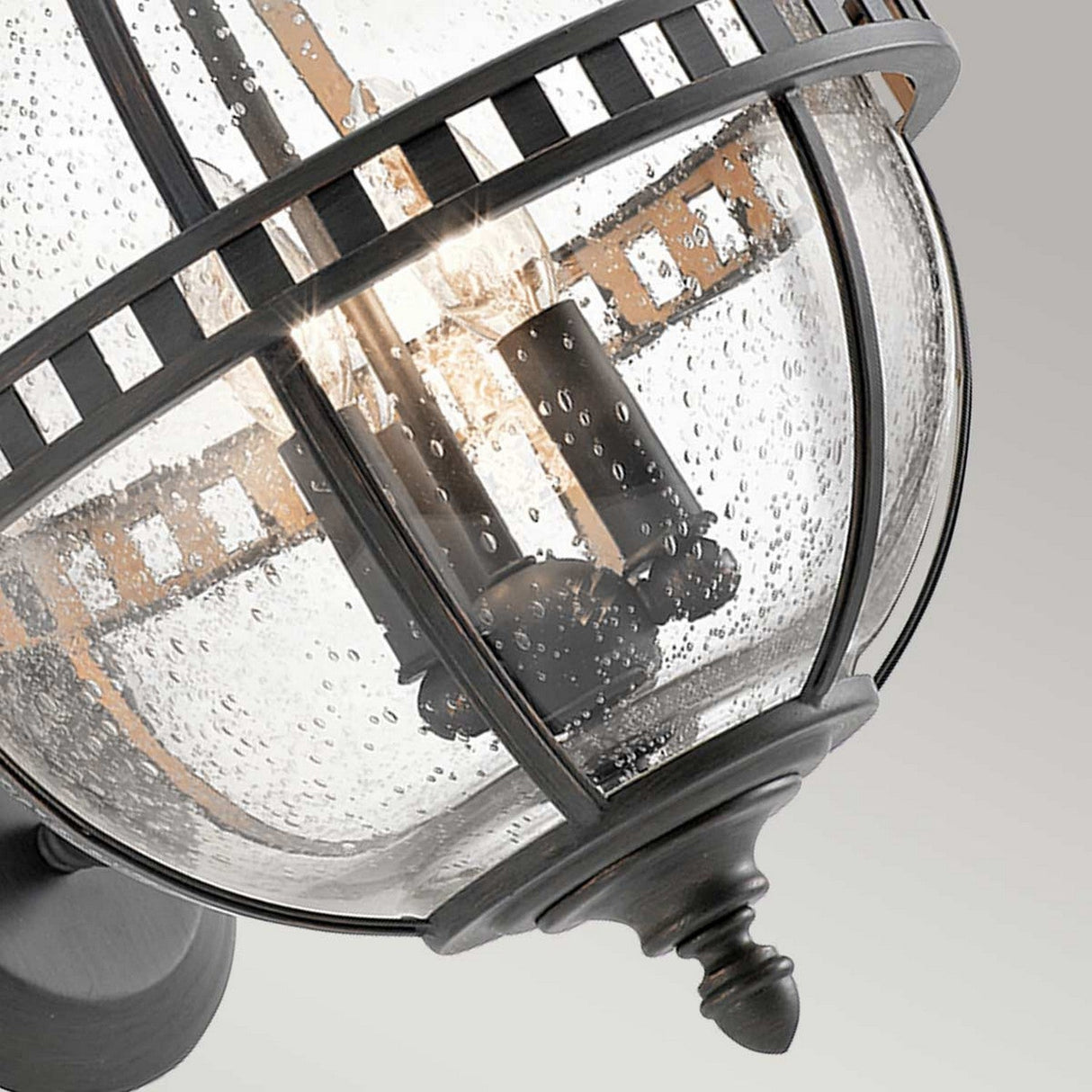 Close-up of the Halleron Outdoor 3 Light Wall Lantern - Grey, showcasing its spherical glass shade and grid-like metal frame. Inside, multiple bulbs are visible through the textured, bubbled glass, casting a warm glow with Victorian sophistication. The background is a plain, soft gray.