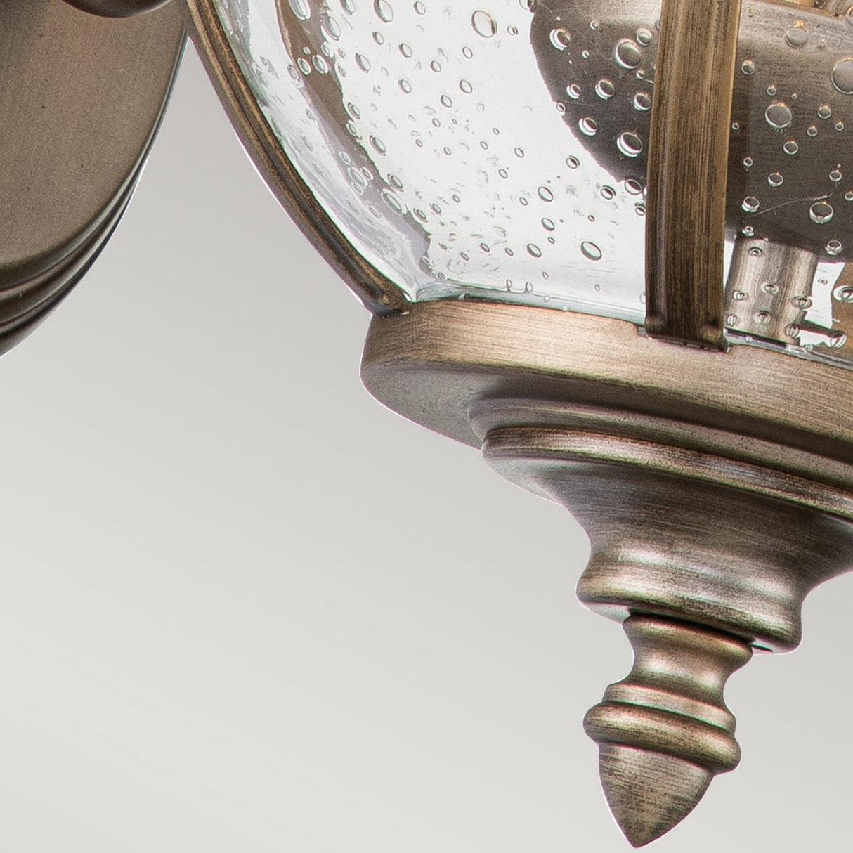 A close-up of the Halleron Outdoor 2 Light Wall Lantern in burnished bronze showcases its textured glass globe with bubble-like patterns reminiscent of Victorian-era lighting. This decorative outdoor fixture features a classic and elegant design, complete with a pointed finial at the base.