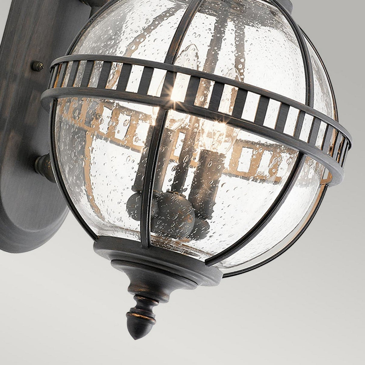 A close-up of the Halleron Outdoor 2 Light Wall Lantern showcases its orb-shaped, clear seeded glass cover encased in a black metal cage design. The illuminated light casts a warm glow, and the fixture exudes Victorian-inspired charm as it is mounted on a dark wall plate.