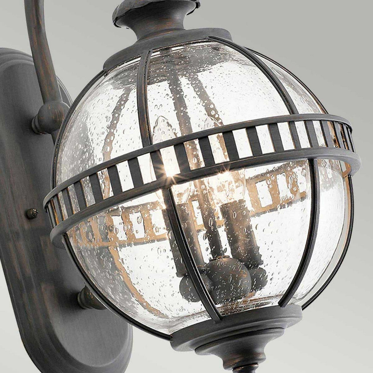 The Halleron Outdoor 2 Light Wall Lantern in Grey offers a close-up view of vintage-inspired charm, featuring its clear seeded glass globe and metal cage-like details. The lights within create a warm glow, while the fixture is elegantly mounted on an oval backplate with a patina finish.
