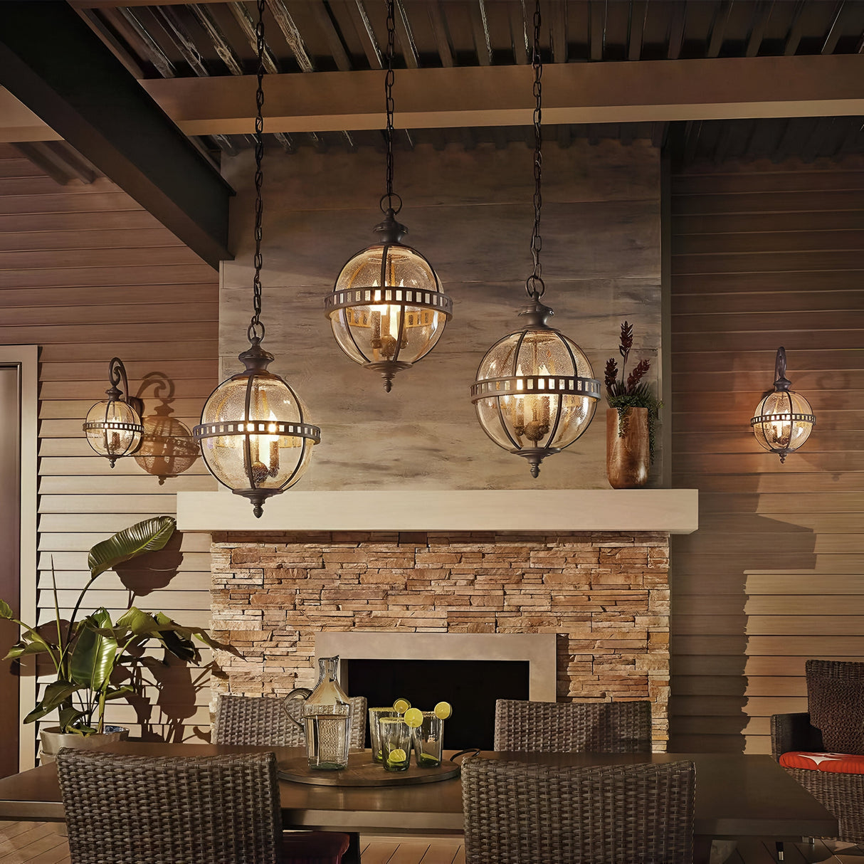 A cozy patio with Victorian-inspired charm features a stone fireplace, surrounded by wicker chairs and a dining table. The space is illuminated by the elegant Halleron Outdoor 2 Light Wall Lantern in grey, which adds warmth with its clear seeded glass. A potted plant on the mantel and lush greenery create a welcoming atmosphere.