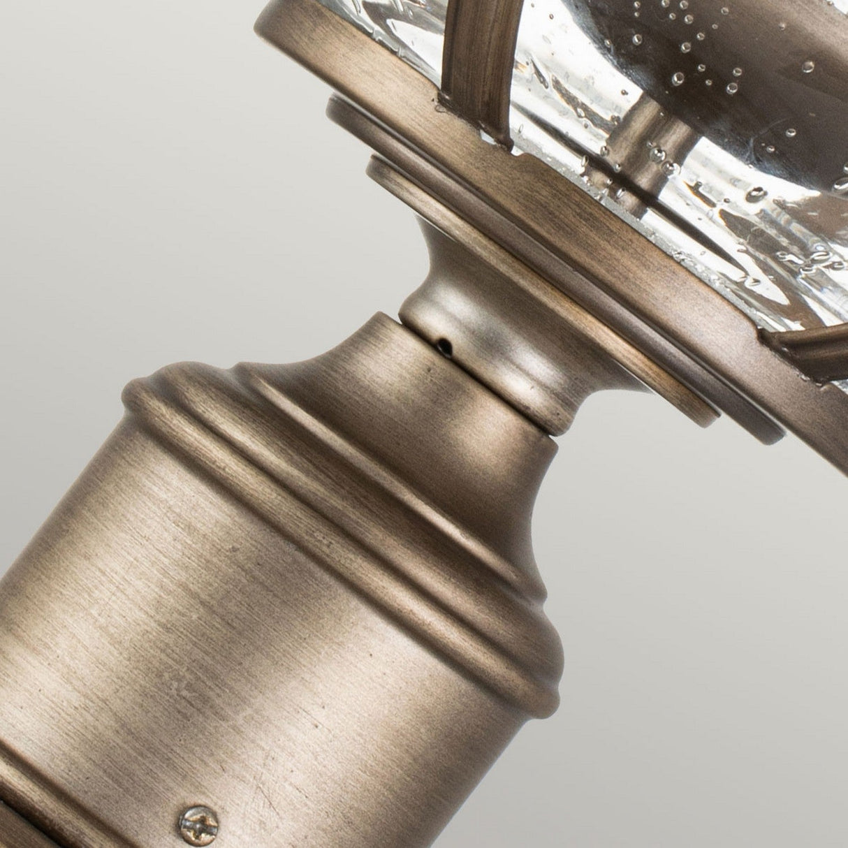 Close-up of the Halleron Outdoor 3 Light Pedestal Pendant in burnished bronze, showcasing a weathered brass finish evocative of Victorian Era lighting. It features clear glass panels with a bubbled texture, mounted on a cylindrical base. The background is light gray.
