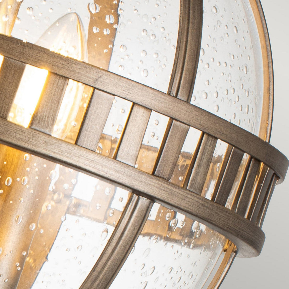 Close-up of the Halleron Outdoor 3 Light Pedestal Pendant in burnished bronze, featuring a spherical design with a warm glowing bulb. The frame showcases vertical and horizontal bars, and clear glass panels adorned with tiny bubbles evoke the elegance of Victorian era lighting.