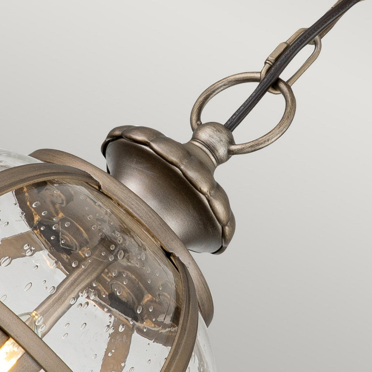 A close-up of the Halleron Outdoor 3 Light Chain Pendant reveals a Victorian-inspired design with a clear glass dome featuring bubble-like details. Its burnished bronze metal casing and chain have a weathered finish, enhancing the lantern's rustic appearance.