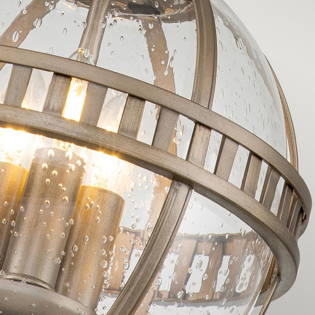 Close-up of the Halleron Outdoor 3 Light Chain Pendant in burnished bronze, featuring a clear, textured glass globe encircled by metal bands. The lighting creates a warm glow that accentuates the bubble-like texture of the glass and the fixture's rich finish.