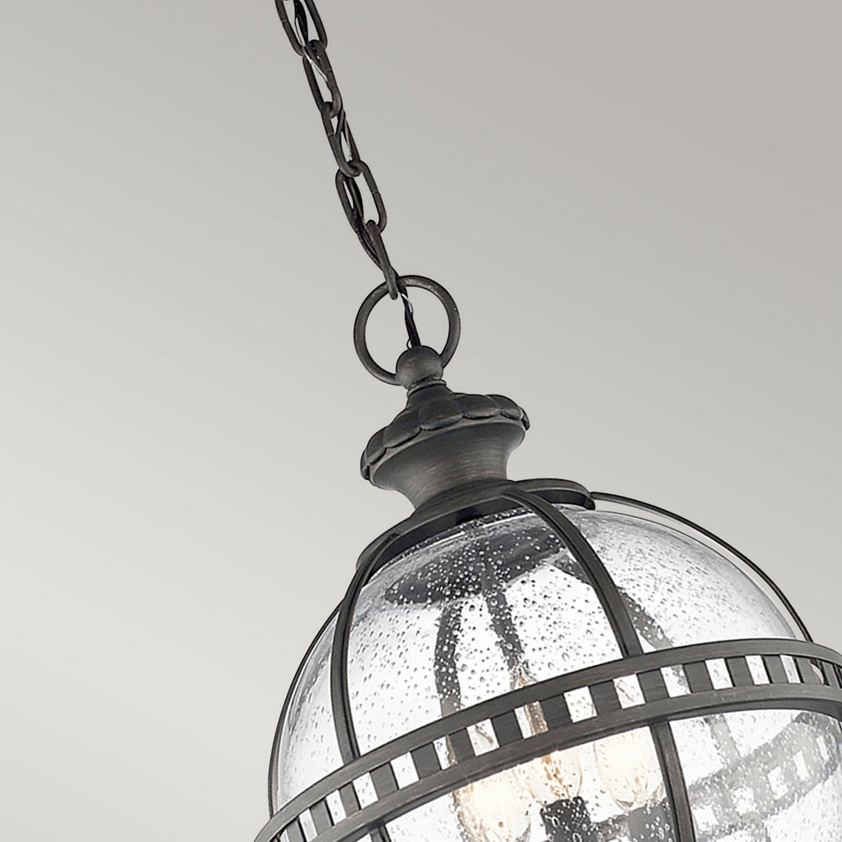 A close-up of the Halleron Outdoor 3 Light Chain Lantern in Grey showcases a vintage-style hanging light fixture with a glass dome and metal frame. The fixture is suspended by a chain, and the glass exhibits a textured, speckled appearance.