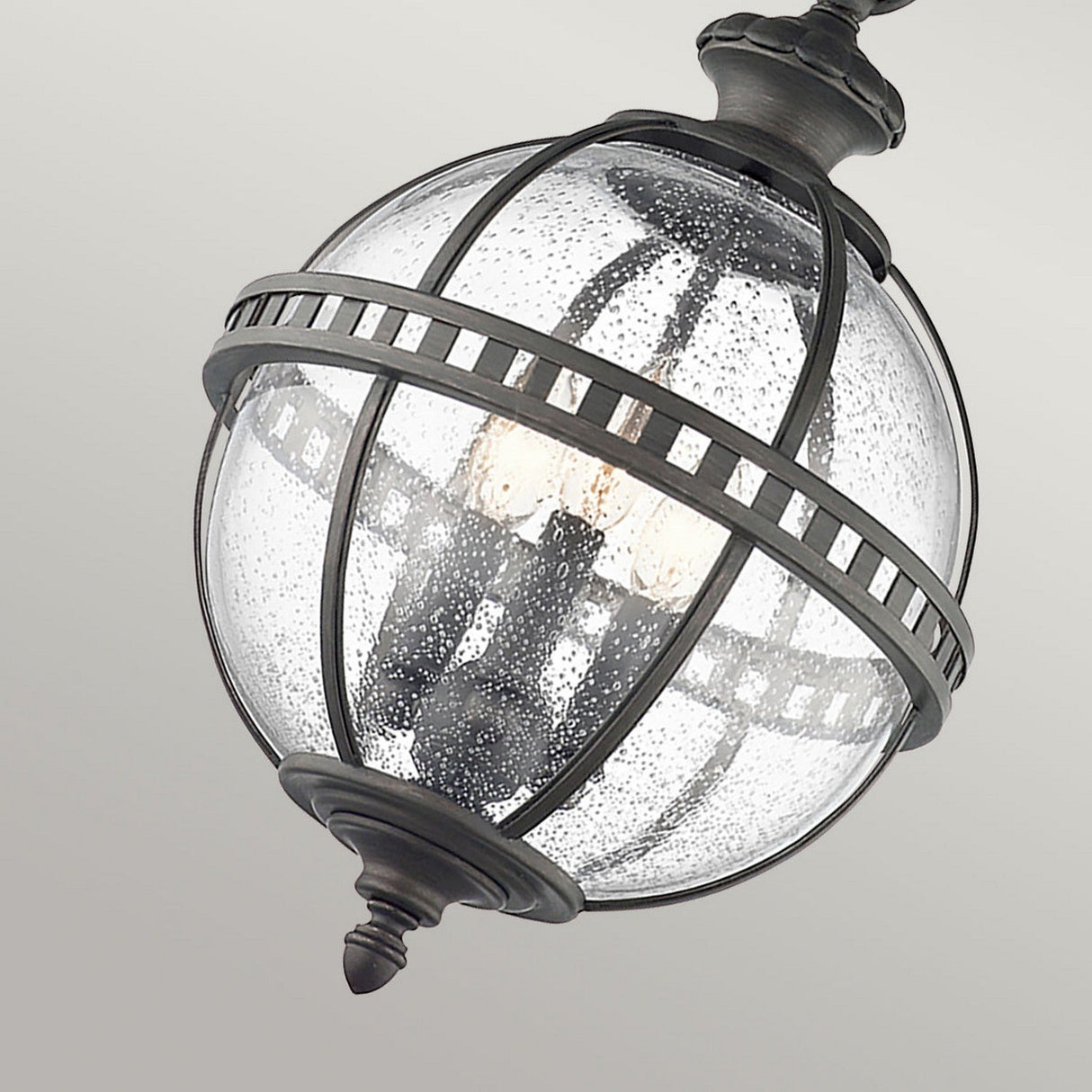 A close-up of the Halleron Outdoor 3 Light Chain Lantern in grey, showcasing its Victorian-inspired design with clear seeded glass and metal bands. Inside, multiple vintage-style light bulbs evoke the charm of a classic chain lantern against a neutral background.