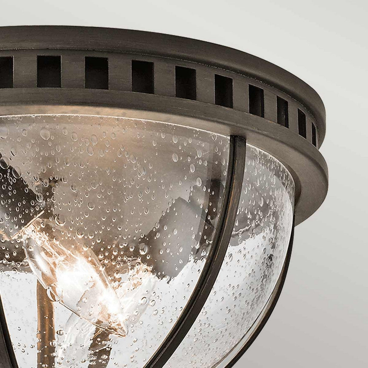 A close-up of the Halleron Outdoor 3 Light Ceiling Flush - Bronze reveals its metal frame paired with a clear, bubbled glass dome that enhances its Victorian design. The bronze finish complements this aesthetic, while inside, three glowing bulbs cast a warm glow from the plain, light-colored ceiling.