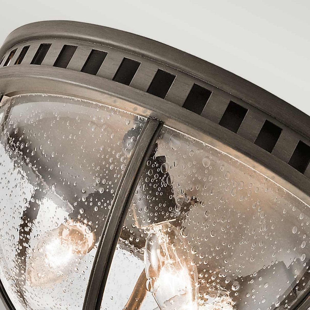 Close-up of the Halleron Outdoor 3 Light Ceiling Flush in a bronze finish, featuring a metal frame and clear, textured dome. This outdoor lighting fixture showcases raindrop-like patterns on the glass with two visible light bulbs inside, emitting a warm glow that evokes Victorian design.
