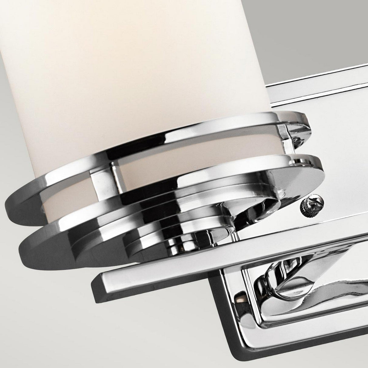 A close-up of the Hendrik 2 Light Wall Light - Polished Chrome, a contemporary silver metallic bathroom fixture, showcases its cylindrical glass encased within horizontal metal rings. Mounted on a glossy rectangular base, the light is turned off against a white background.