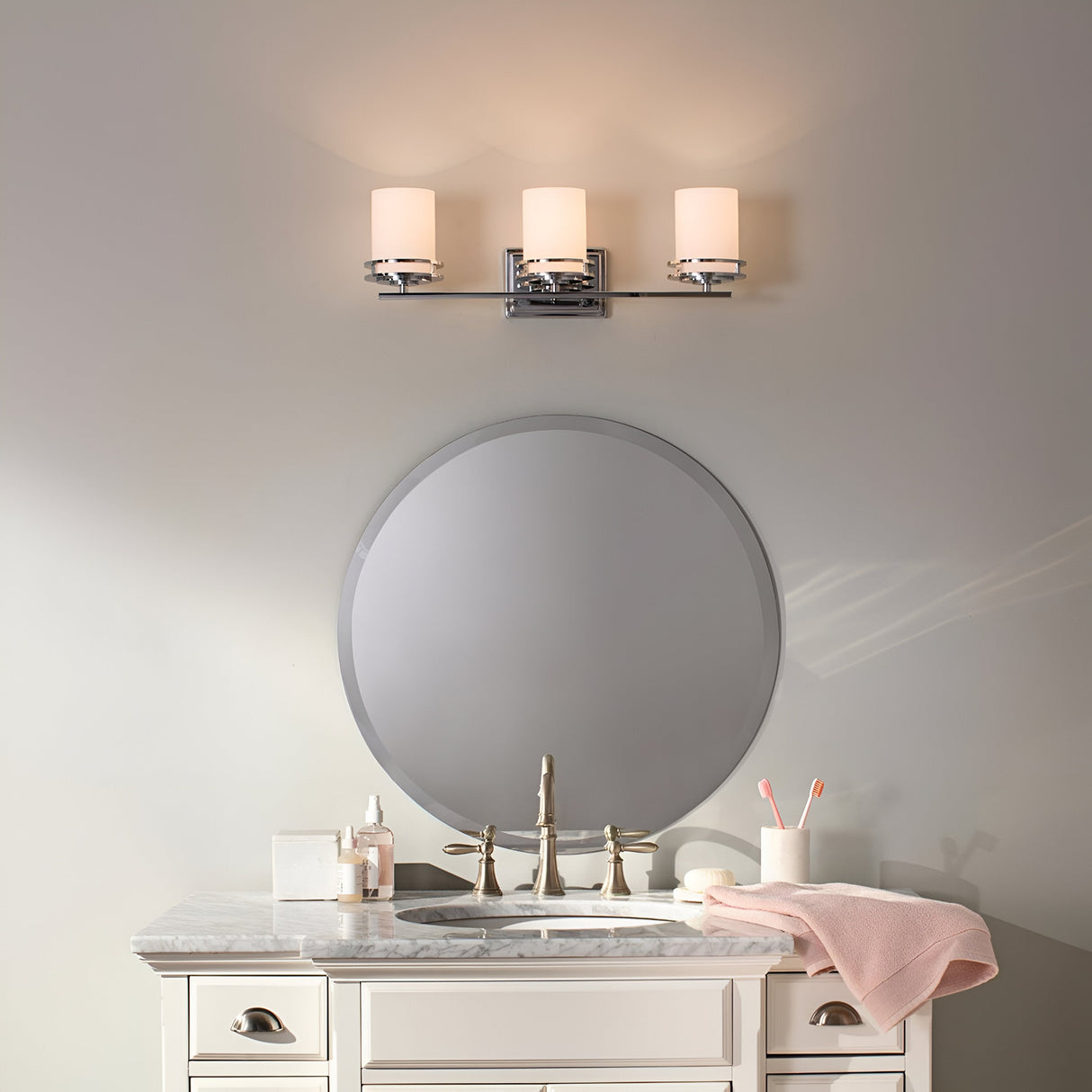 A bathroom features a round mirror above a marble countertop with a beige vanity. The sink faucet is centrally placed, accompanied by a pink towel and soap dispenser on the counter. For lighting, the room boasts the Hendrik 3 Light Wall Light in polished chrome, which includes three candle-shaped lights offering both style and function.