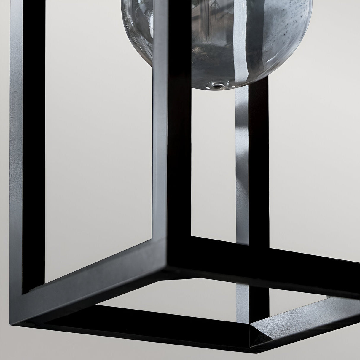 A close-up of the Hone Coastal 1 Light Medium Outdoor Wall Lantern in black, featuring a minimalist geometric metal frame and a glass bulb, set against a neutral, light background.
