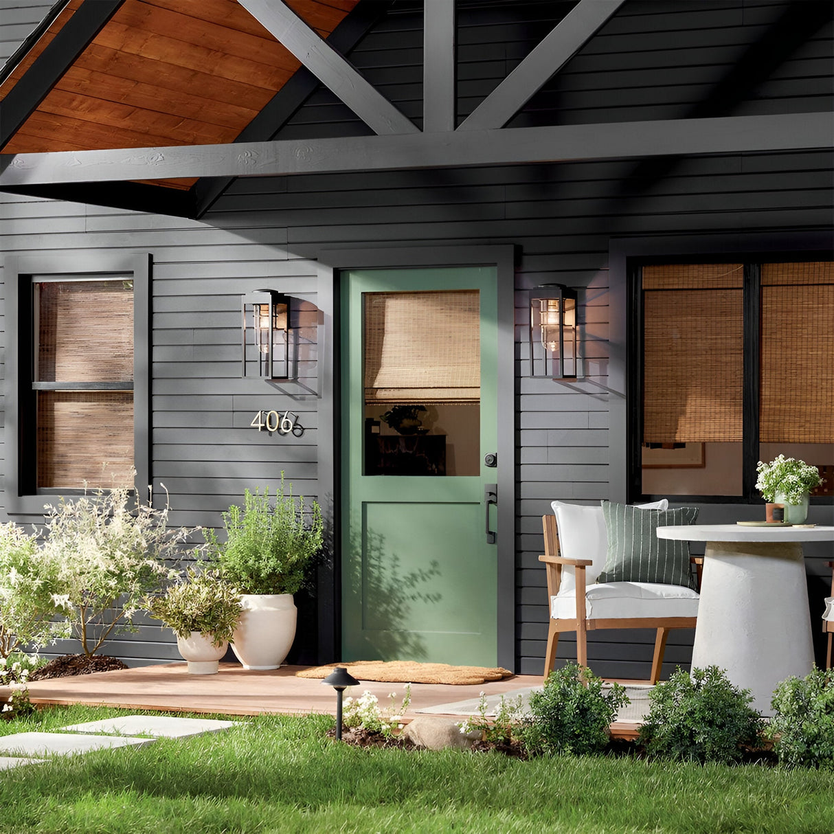 A modern house exterior with dark gray siding, a green front door, and large potted plants. The Hone Coastal 1 Light Medium Outdoor Wall Lantern in black brightens the porch, which features a white chair, table, and house number 4066.