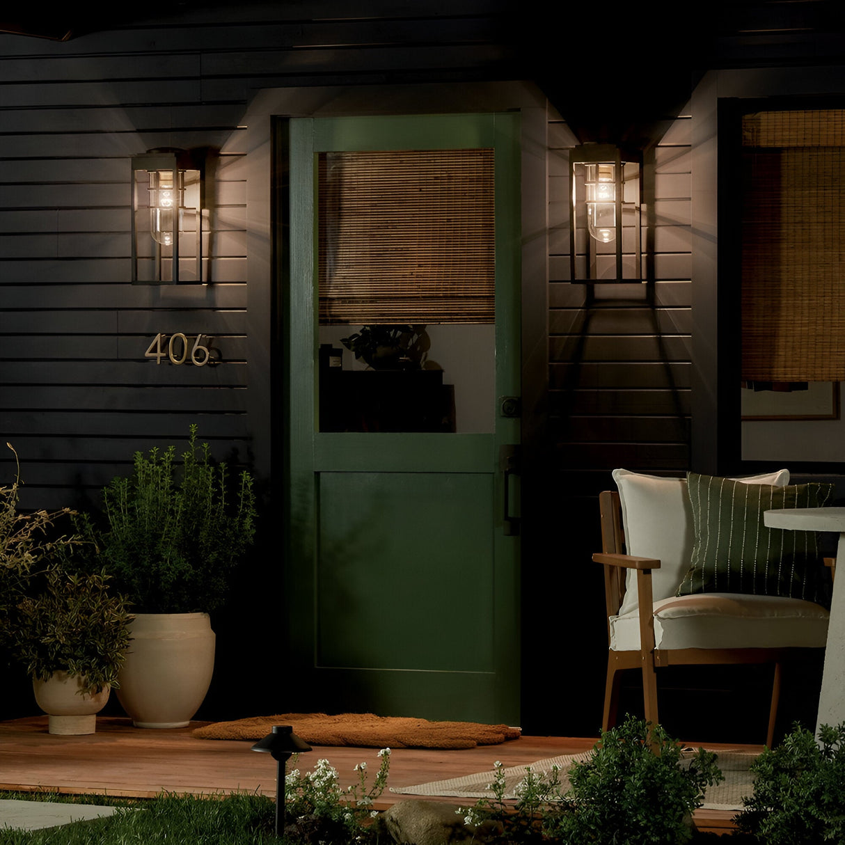 A green door with a window and bamboo shade is flanked by Hone Coastal 1 Light Medium Outdoor Wall Lanterns in black. A welcome mat, potted plants, and a cushioned chair with pillows complete the cozy porch setting for evening relaxation.