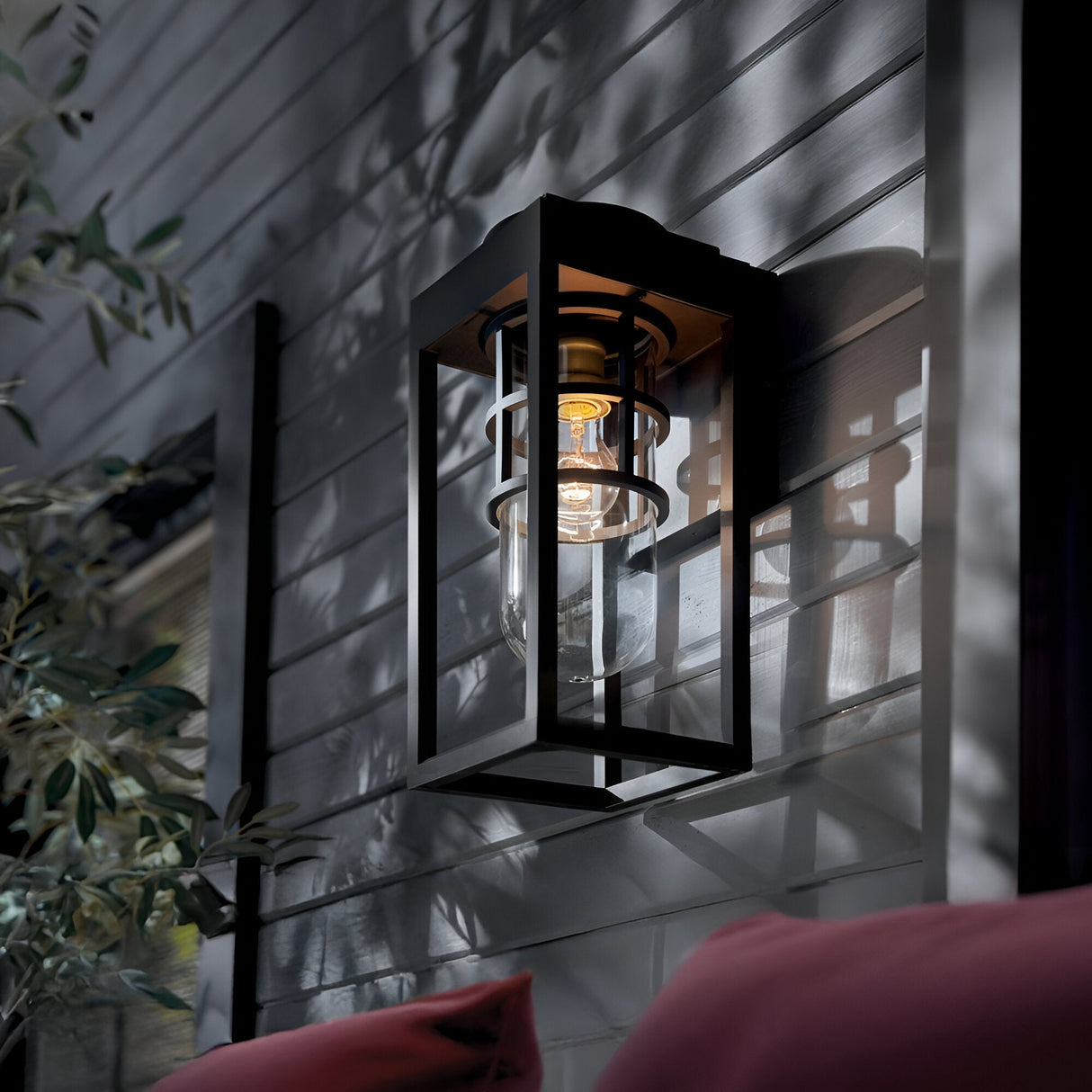 The Hone Coastal 1 Light Small Outdoor Wall Lantern in Black features a weather-resistant design and clear glass bulb, glowing warmly at dusk against a gray wall with leafy branches and red-cushioned furniture nearby.