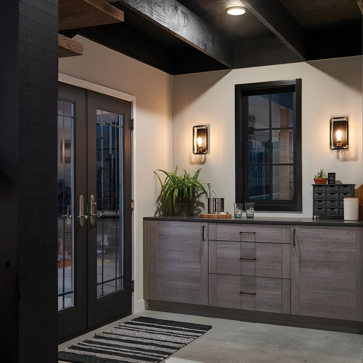 A modern, minimalist interior features gray cabinets, a glass door, and a window. The cabinet displays a potted plant and various decor items. Two Imahn 1 Light Wall Lights in black and natural brass with Edison-style bulbs illuminate the space, while a striped rug covers the floor. Dark wooden beams accentuate the industrial design of the ceiling.