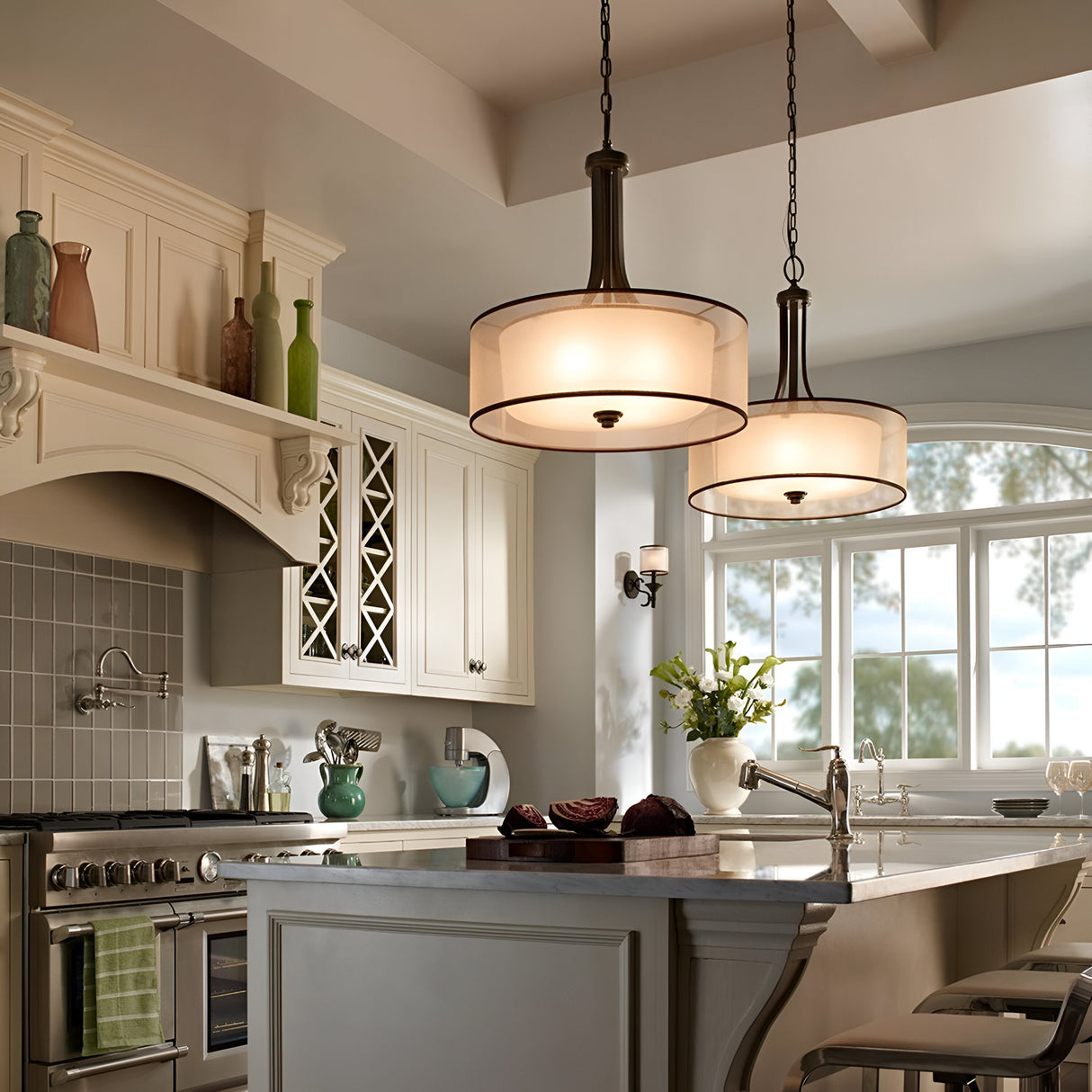 A bright kitchen featuring the elegant Lacey 4 Light Large Pendant in Bronze, with its sophisticated double-shade design, hangs above a central island equipped with a sink and modern appliances. Cabinets and shelves display decorative items, while a large window floods the space with natural light.
