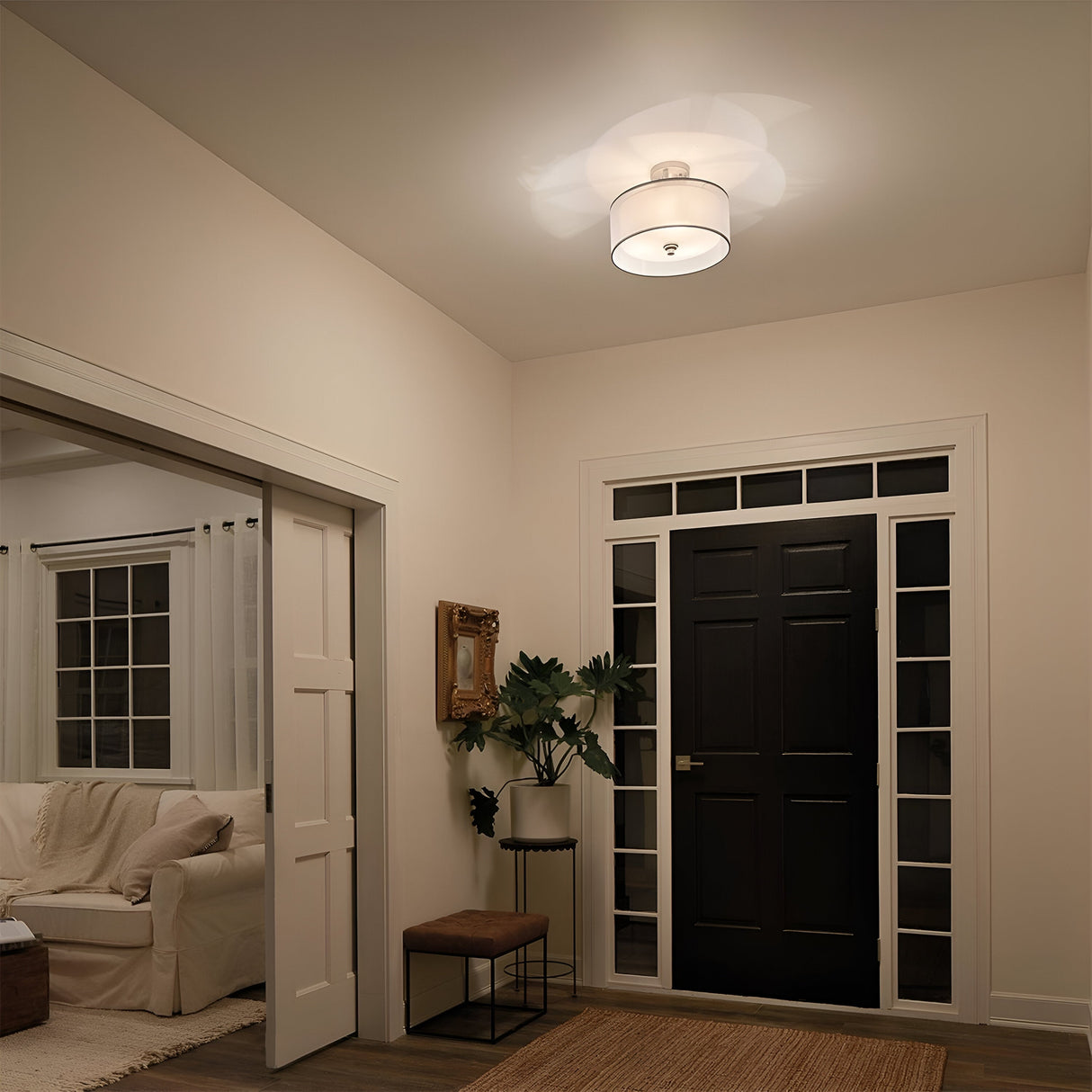 A well-lit entryway features a black front door with glass panes, a small mirror, a plant, and a cushioned stool. The adjacent living room is visible through an open doorway, displaying a white sofa and a small side table. The Lacey 3 Light Small Semi Flush in polished silver casts a layered glow across the space.