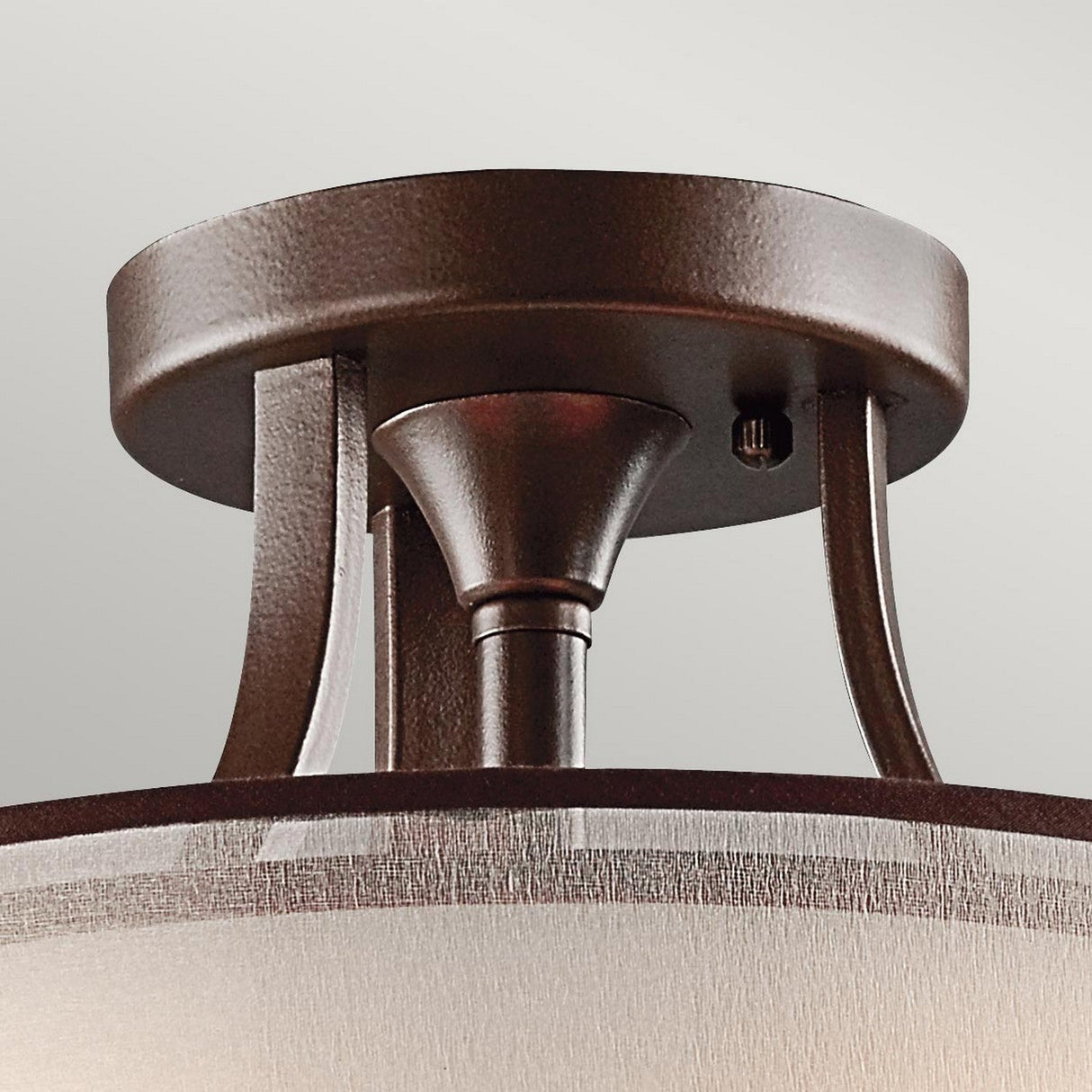 The Lacey 3 Light Small Semi Flush in bronze showcases a modern yet simple design with curved metal supports. Its round base and cylindrical shade offer a sleek look, elegantly displayed against a plain, light-colored wall.