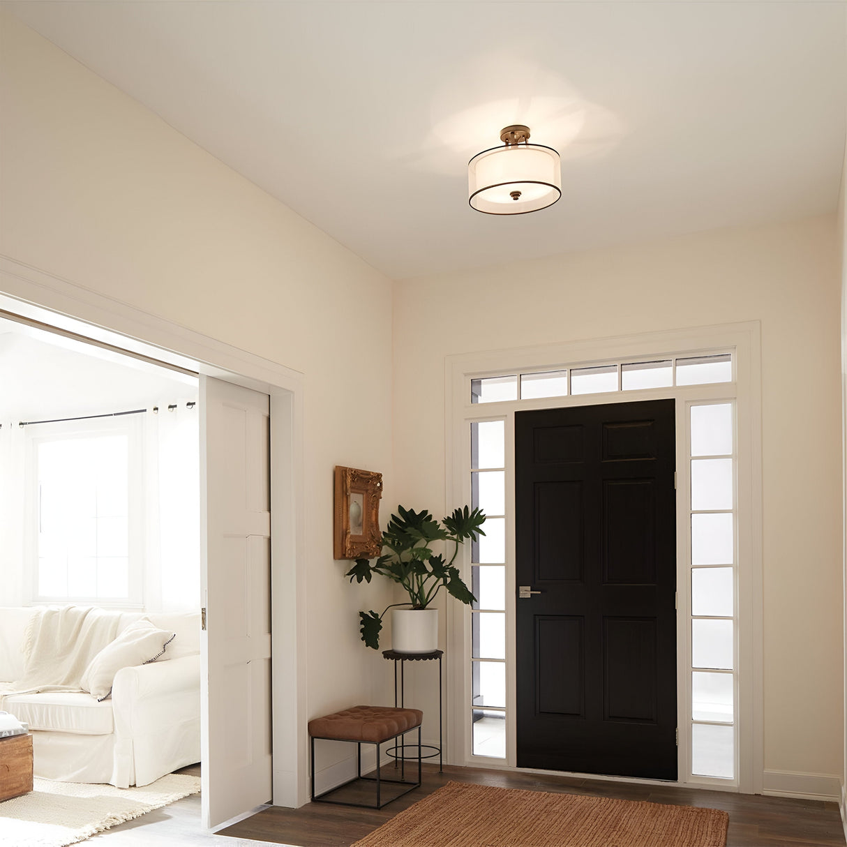 A cozy entryway features a black front door framed by glass panels. A small bench with a cushion sits next to a potted plant and a mirror on the wall. The space is softly lit by the Lacey 3 Light Small Semi Flush - Bronze ceiling fixture, offering a glimpse of the bright living room beyond.