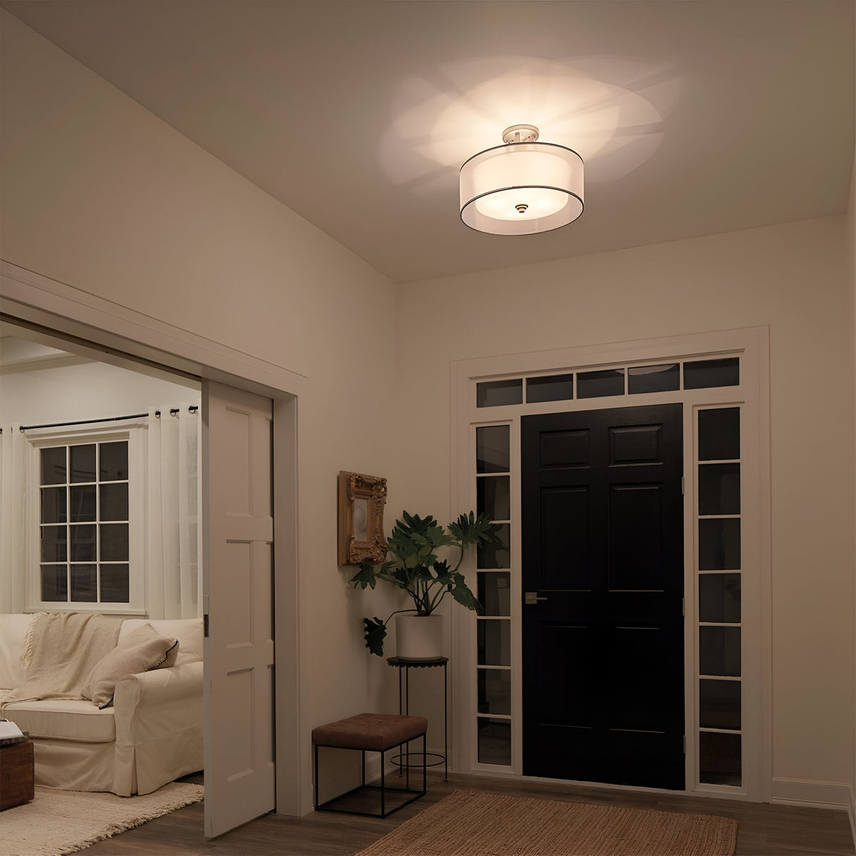 A modern entryway showcases a black front door framed by tall windows. The Lacey 4 Light Semi Flush in a polished silver finish adds an elegant touch of illumination to the space. A plant and small stool are positioned near the door, while a gold-framed mirror adorns the wall. The adjoining room includes a white sofa tastefully adorned with blankets.
