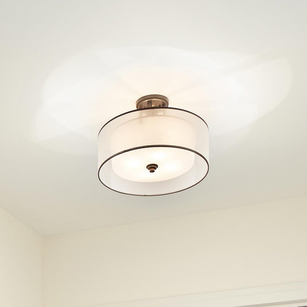 The Lacey 4 Light Semi Flush - Bronze is a contemporary ceiling light fixture that boasts a bronze semi flush design paired with a circular white fabric shade. It casts a soft glow on the beige ceiling and walls, enhancing the elegance of the space with its understated charm.