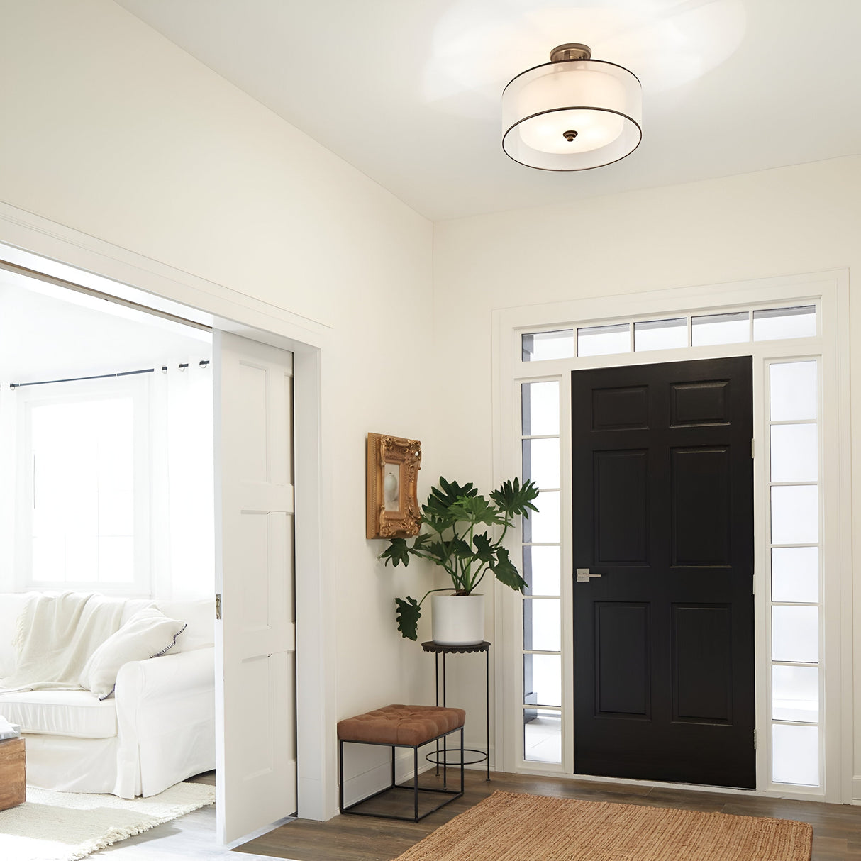 A contemporary entryway with a black front door surrounded by glass panels is warmly illuminated by the Lacey 4 Light Semi Flush in Bronze. A potted plant rests on a small table next to a cushioned stool and square mirror. Through the open sliding door, a white room invites you beyond the beige rug.