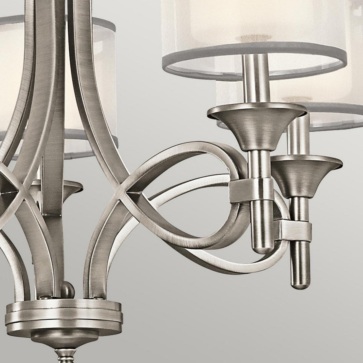The Lacey 5 Light Chandelier - Silver showcases a close-up of its elegant design with metal curves and cylindrical lampshades. This sophisticated lighting piece features intricate metallic loops and smooth, brushed finishes, presenting a sleek look against a light gray background.