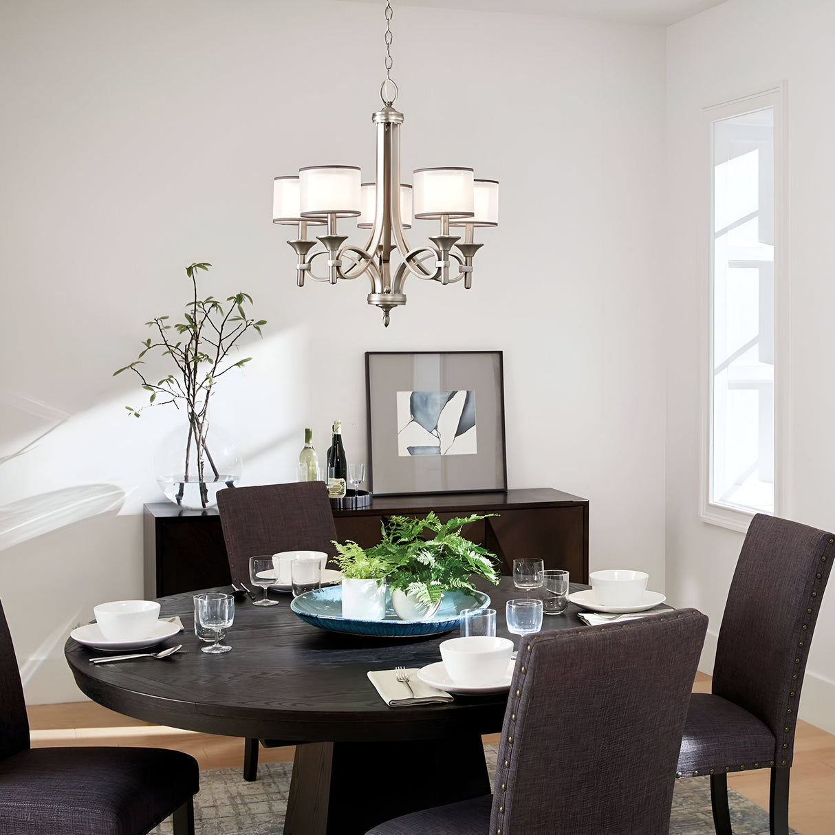 A modern dining room showcases a dark round table set for four, adorned with white bowls, glasses, and a centerpiece featuring greenery. Overhead hangs the elegant Lacey 5 Light Chandelier in silver, while a sideboard with decorative items is positioned against the wall, which is decorated with art and complemented by a nearby window.