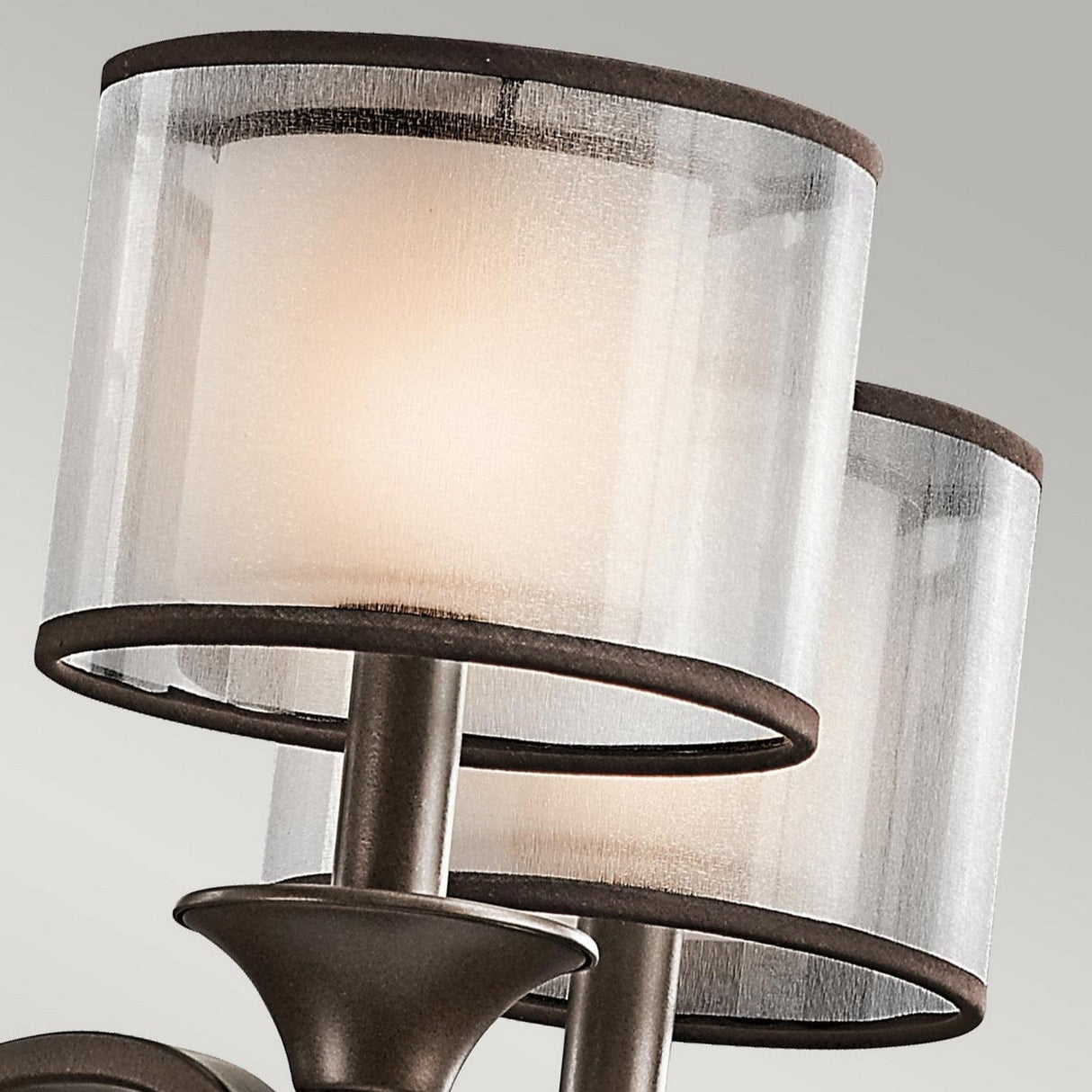 A detailed look at the Lacey 5 Light Chandelier - Bronze reveals its modern design featuring three cylindrical lampshades. These shades emit a warm glow and are transparent with subtle bronze accents that perfectly enhance their metallic fixture, making it ideal for transitional interiors.