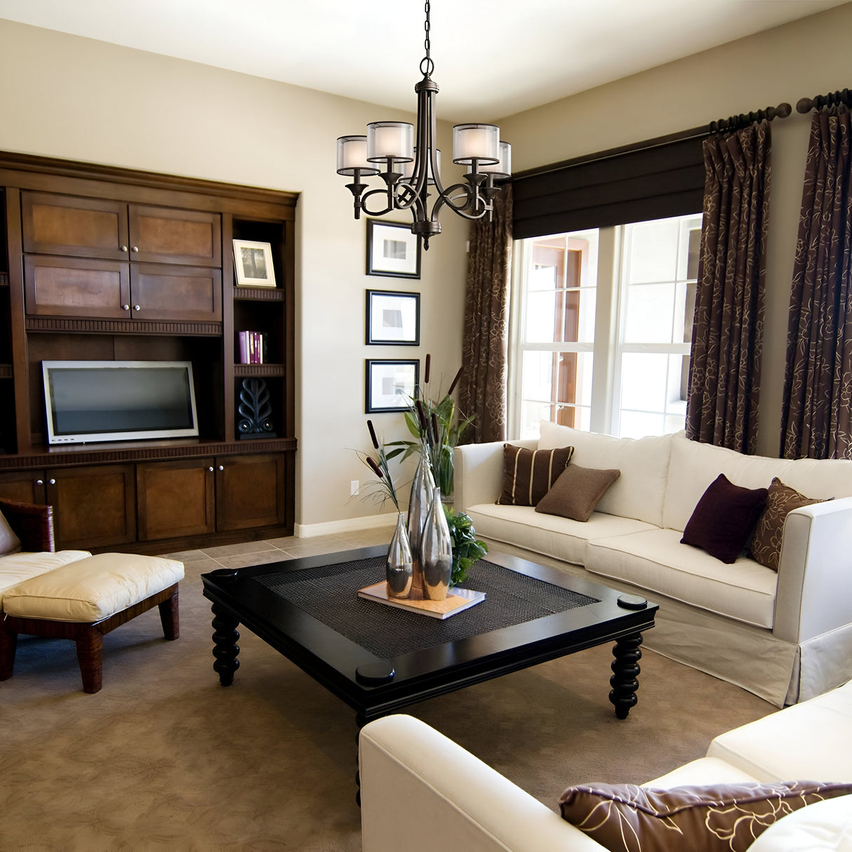 In a cozy living room, a spacious white sofa and a beige armchair surround a dark wooden coffee table. Overhead, the Lacey 5 Light Chandelier in bronze with its distinctive double-shade design illuminates the space. A wooden entertainment center with a TV is positioned against the wall while sunlight pours through the window.